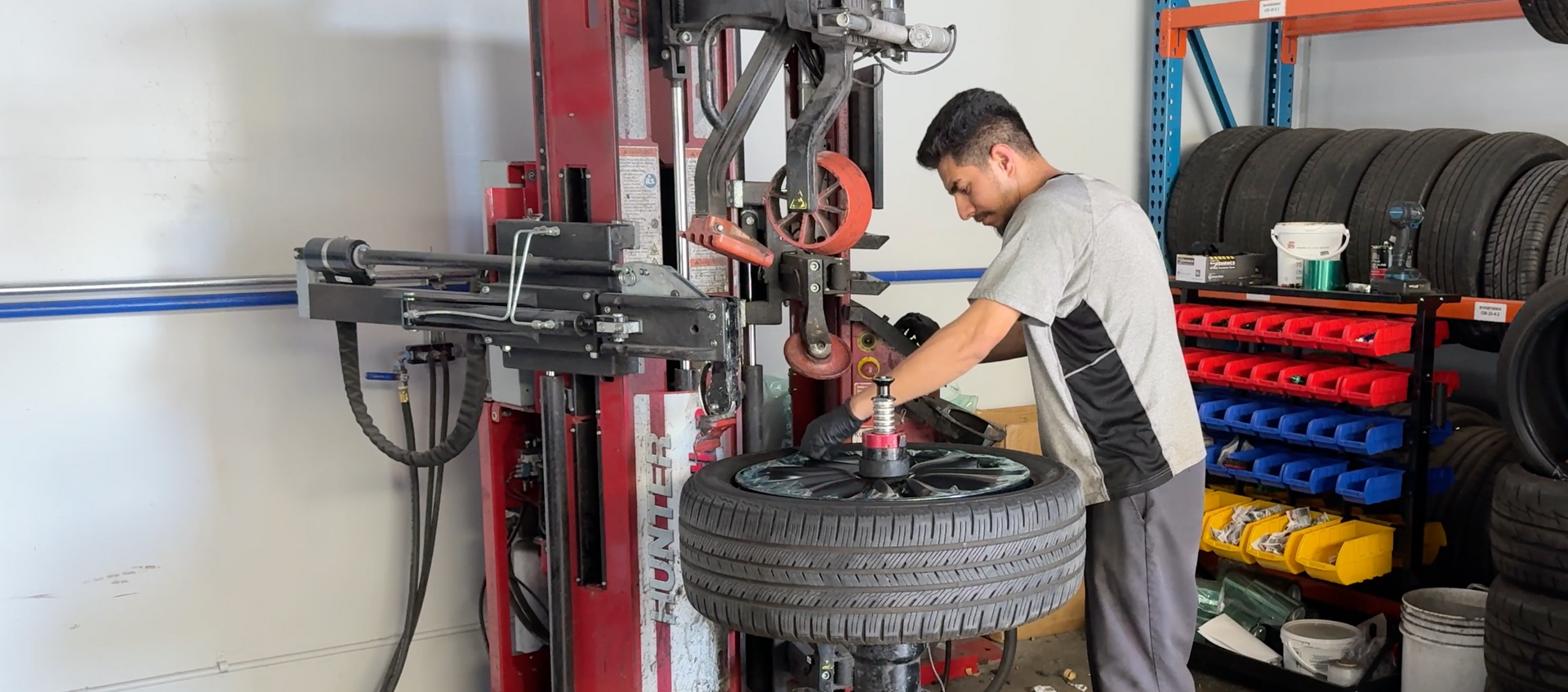 Mechanic working on a tire-changing machine in an auto shop, with stacked tires nearby.