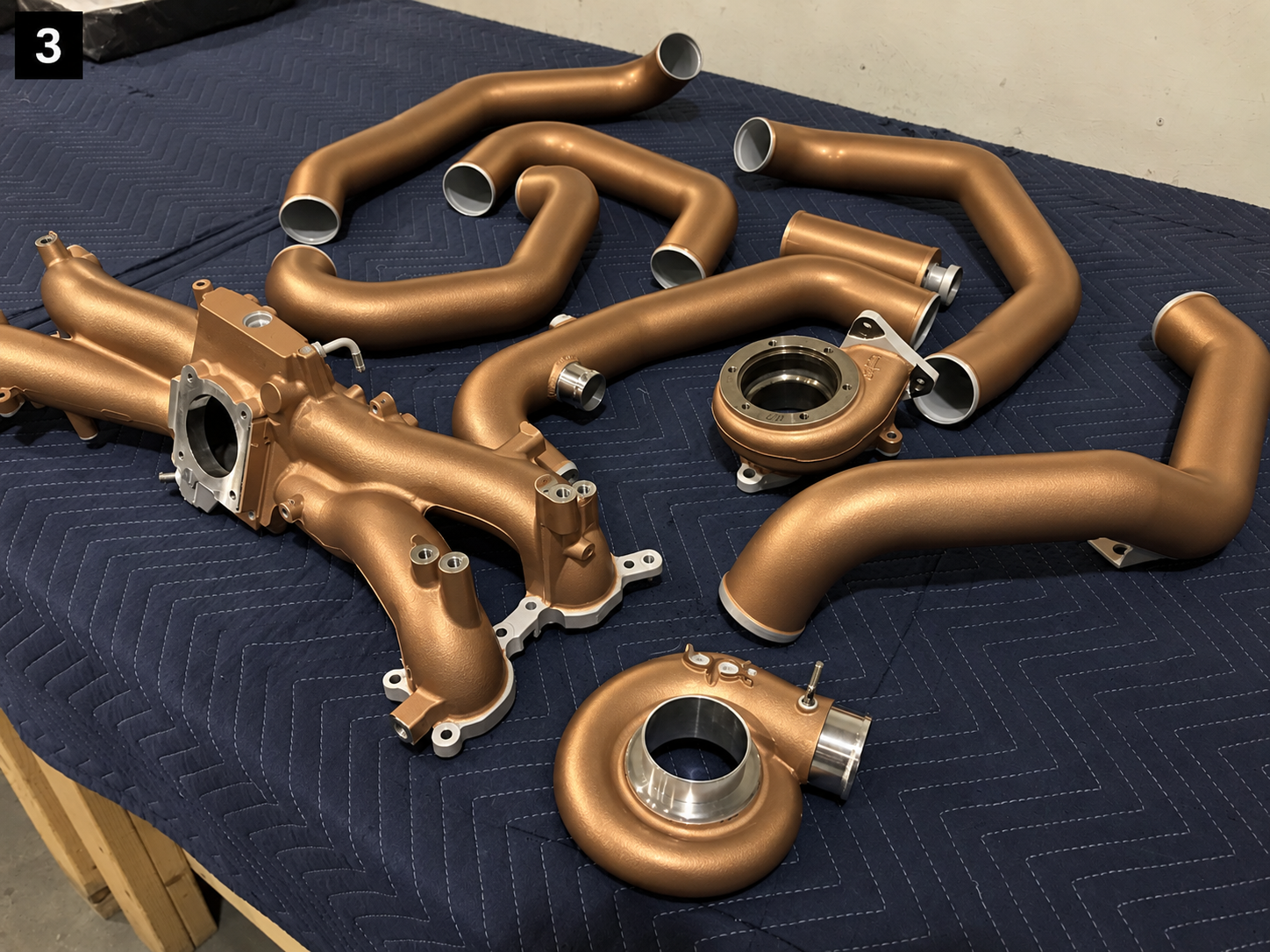 Bronze-colored automotive exhaust pipes and turbo manifolds arranged on a blue quilted workbench.