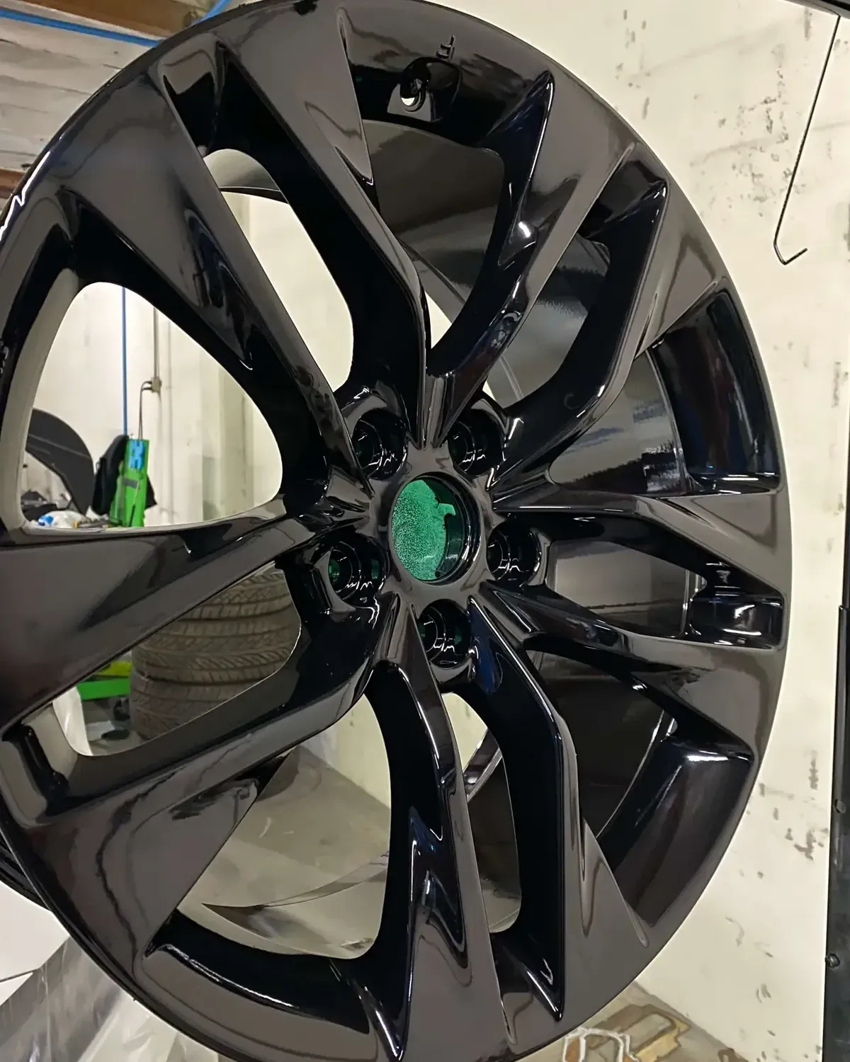 Glossy black car alloy wheel rim with a green center cap, photographed indoors against a white wall.