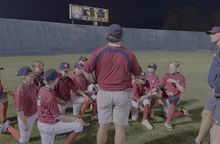 Coach kneels with children on a grassy field during an outdoor sports activity, all gathered in a circle