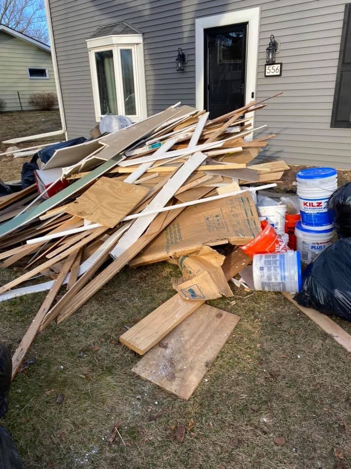 Pile of construction debris and lumber on a lawn beside a gray house with a front door and buckets nearby
