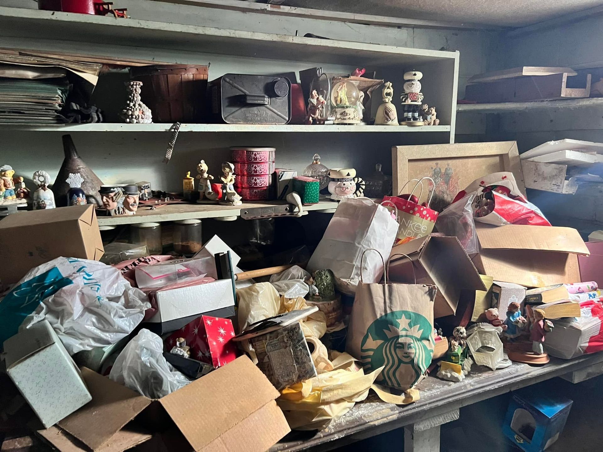 Cluttered shelves and table piled with boxes, bags, and assorted household items in a storage room