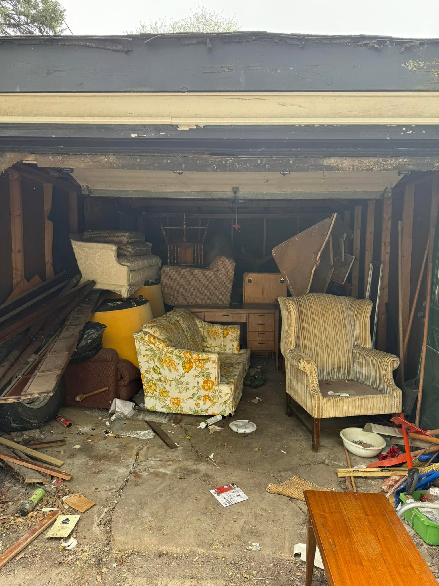 Open garage packed with assorted furniture, boxes, and debris, including two armchairs in front.