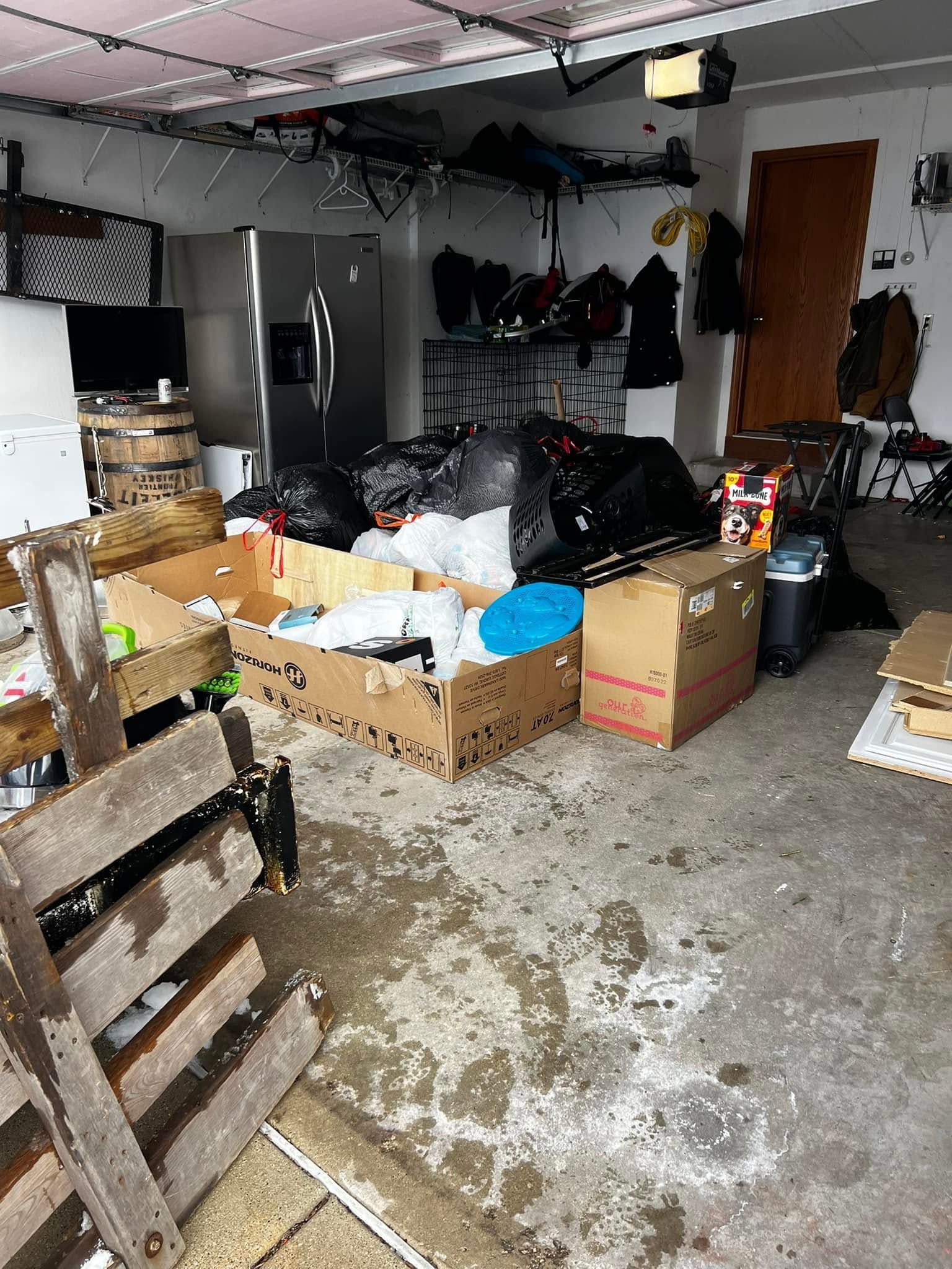 Cluttered garage with stacked boxes, bags, shelves, and a wet concrete floor