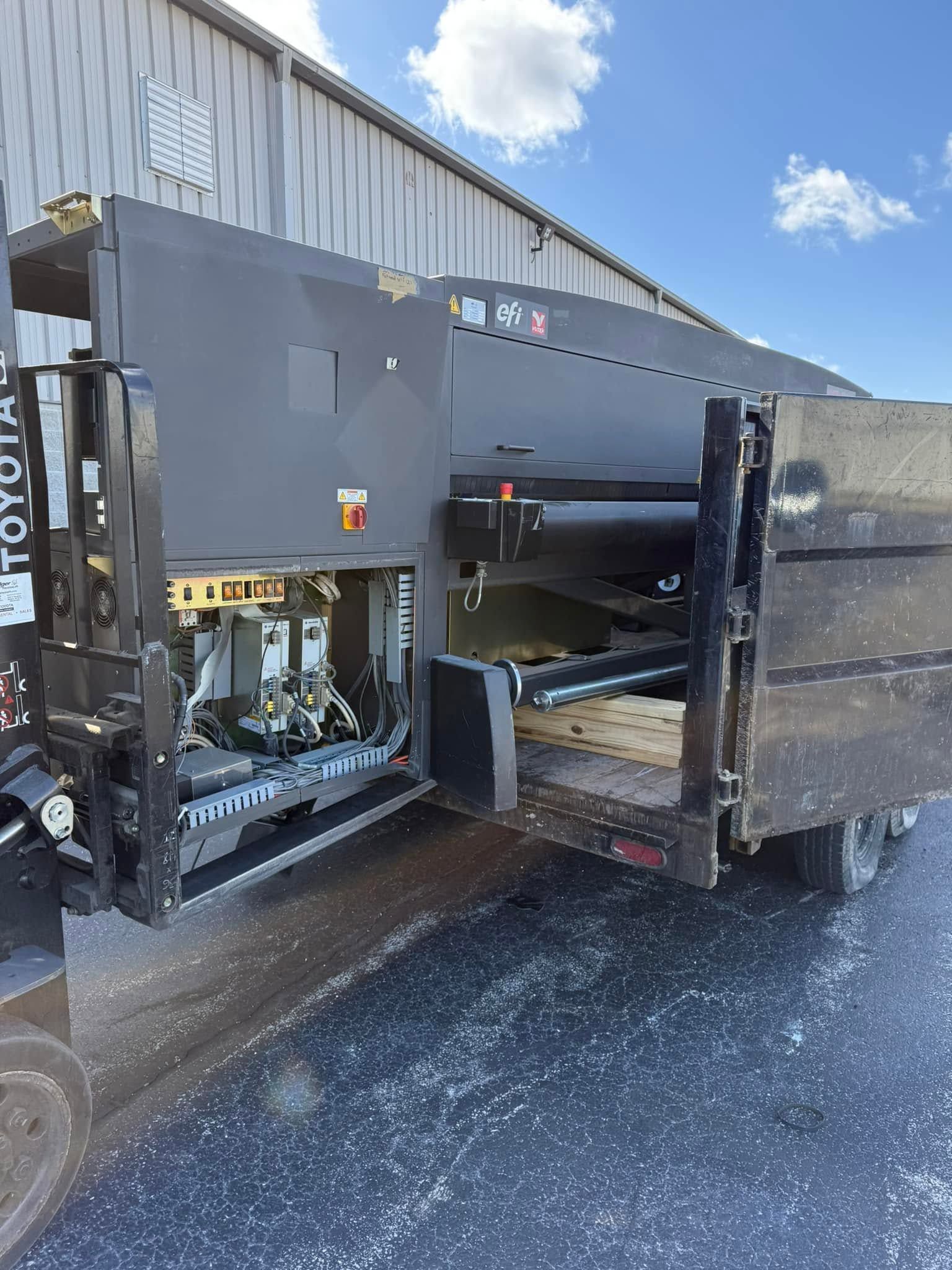 Forklift loading a large gray tank onto a trailer beside a warehouse.
