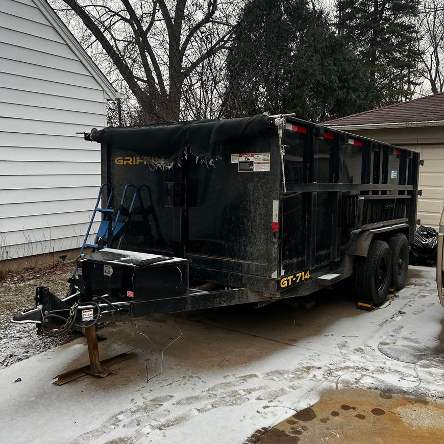 Black dump trailer parked beside a white house on a snowy driveway, with a trailer jack at the front.