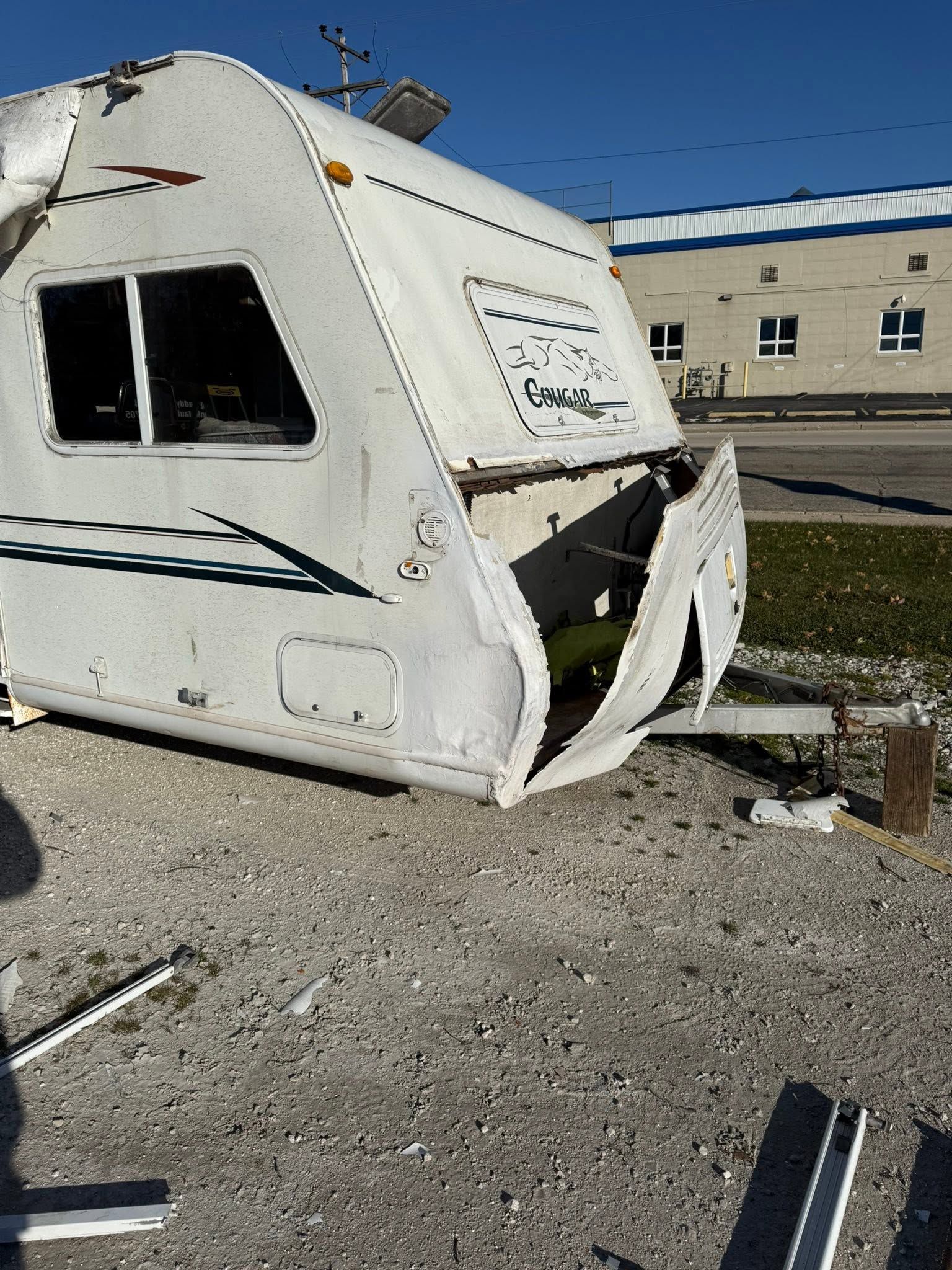 Damaged white camper trailer with a torn front end beside a building and debris on the ground