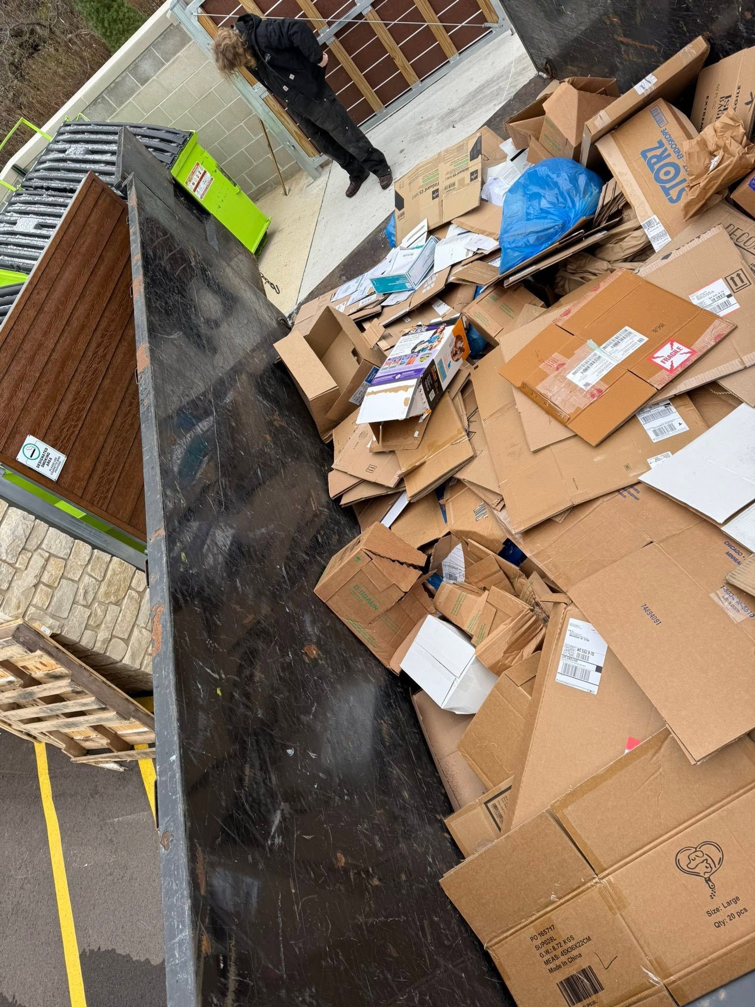 Overflowing cardboard trash in an alley beside a brick building, with a person in dark clothing near the bins