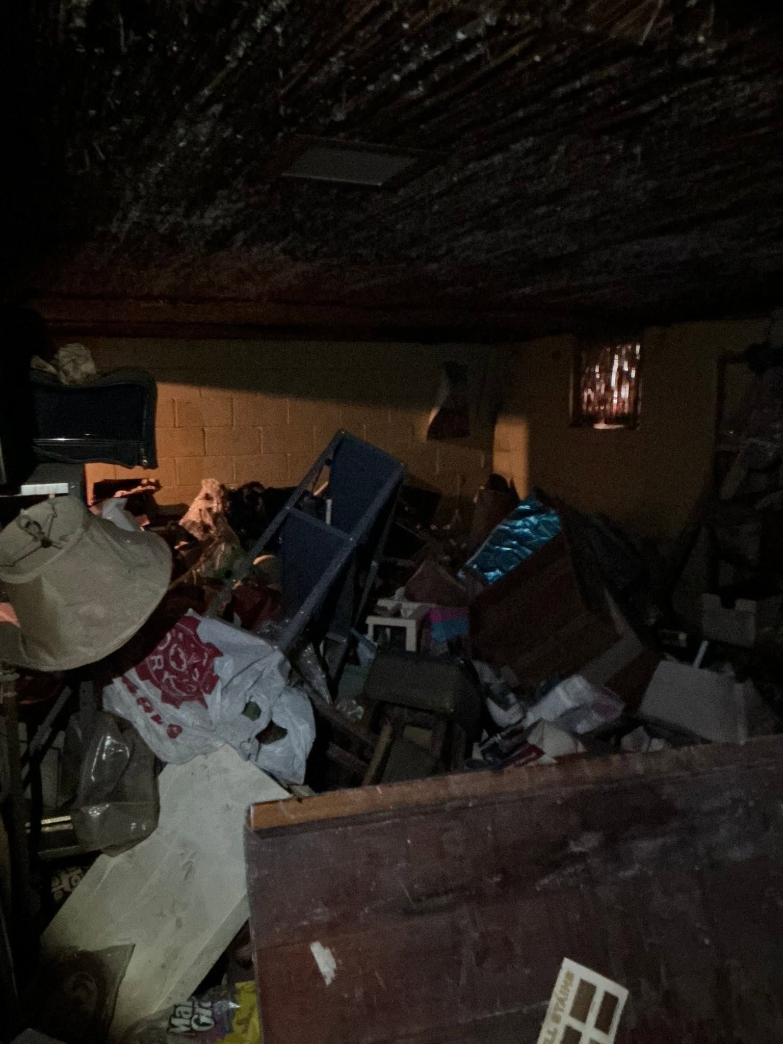 Cluttered room filled with debris and broken furniture, with a damaged ceiling and dim lighting.