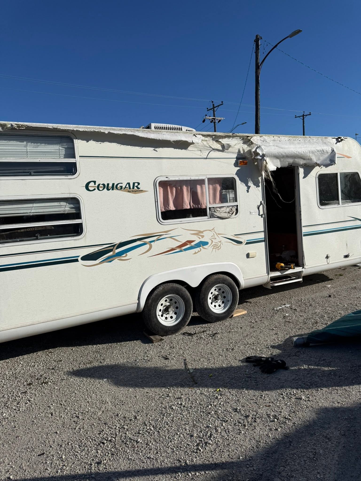White travel trailer with an open door and striped awning parked on a gravel lot under a clear blue sky