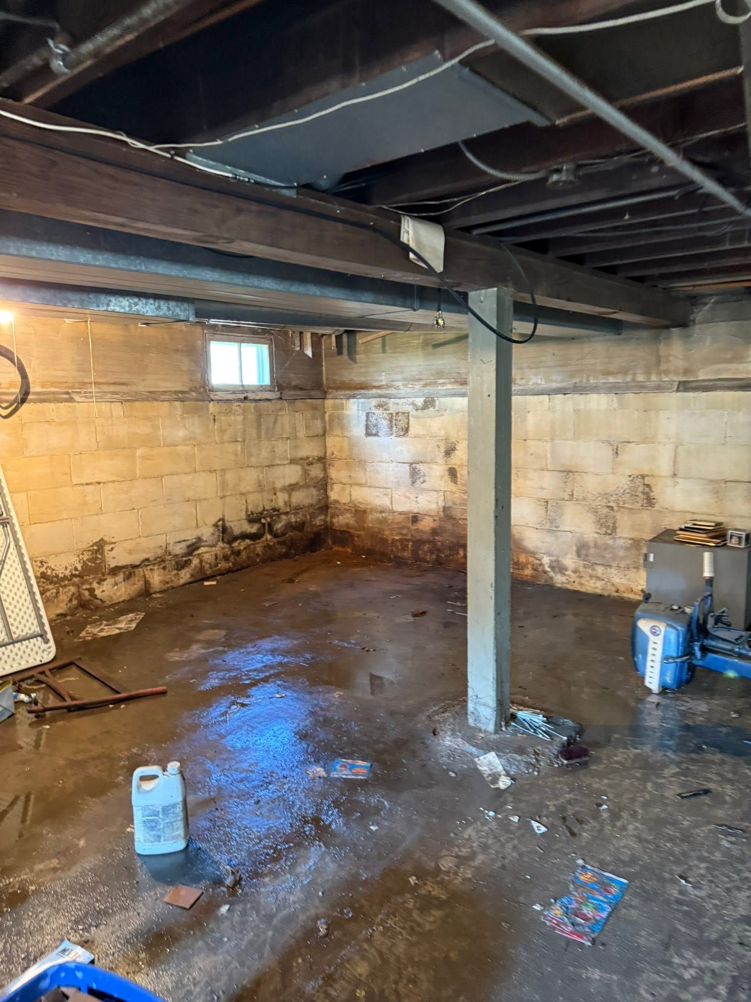Unfinished basement with exposed beams, concrete walls, and a central support pole; wet floor and scattered items.
