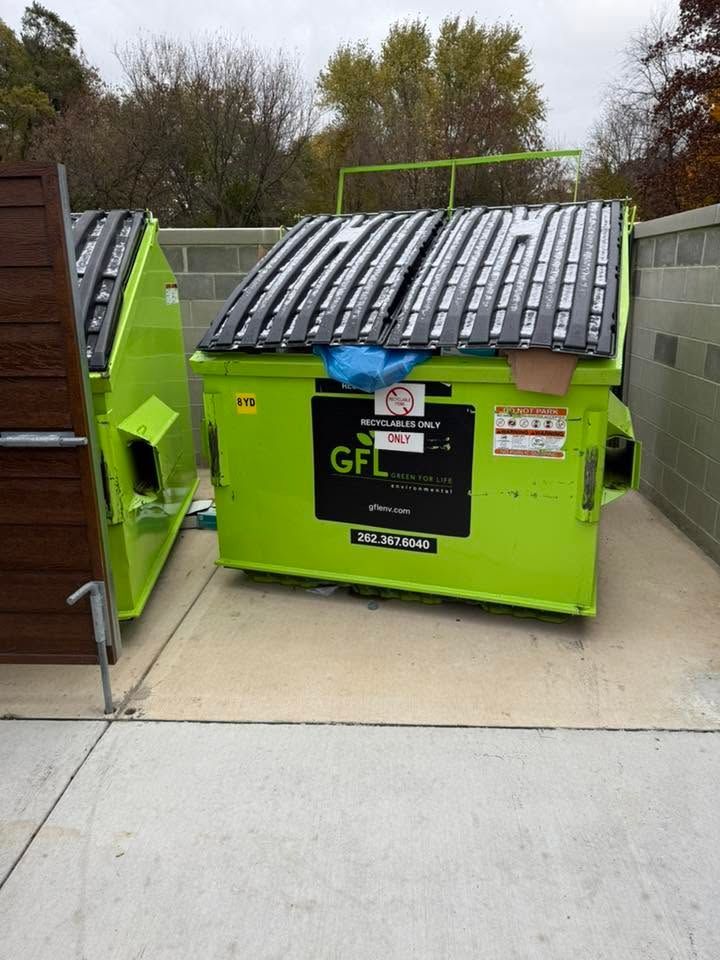 Bright green dumpsters with black lids and recycling signs on a paved area outdoors