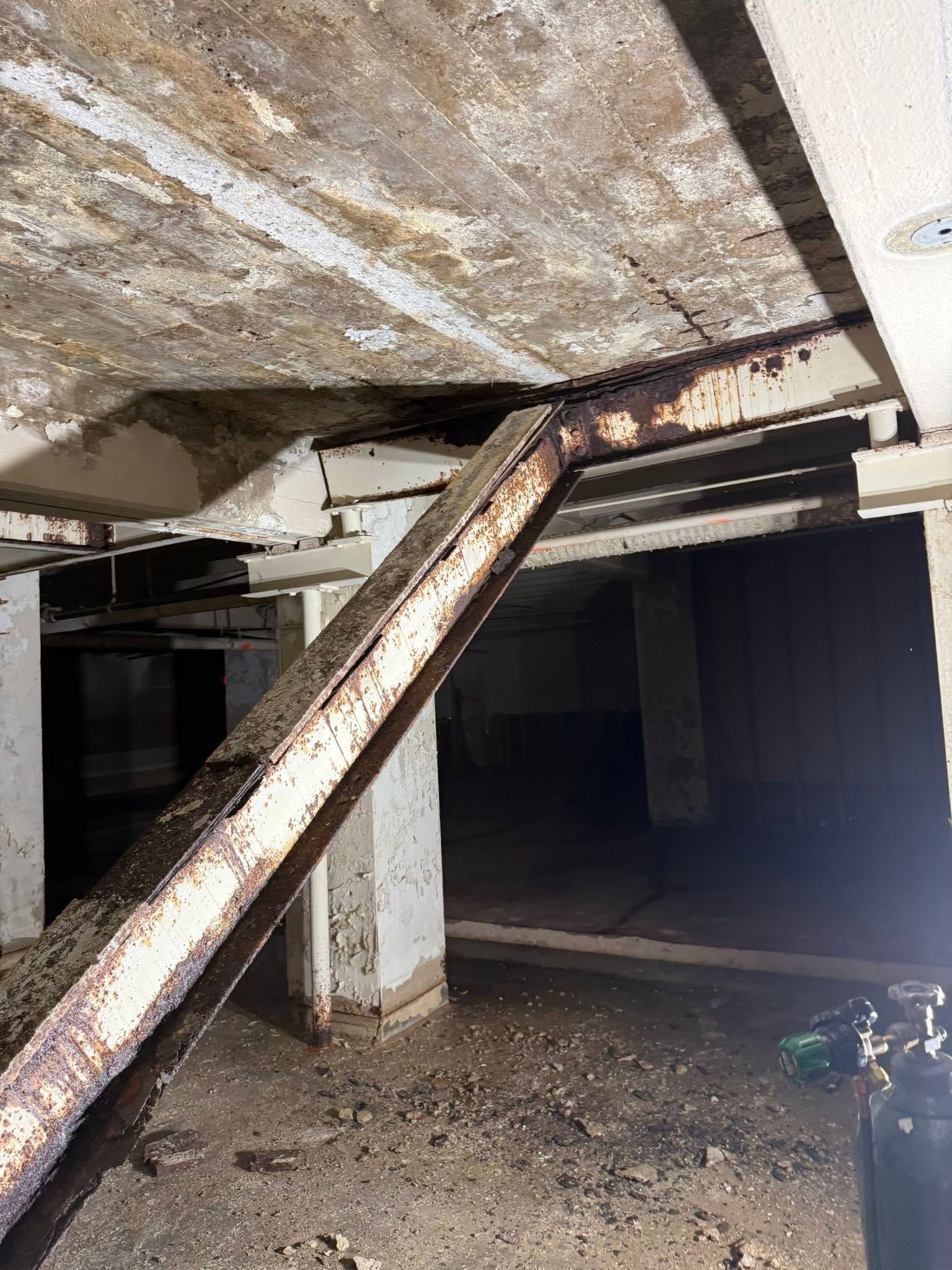 Rusty support beam under a concrete ceiling in a dark crawlspace with dirt floor