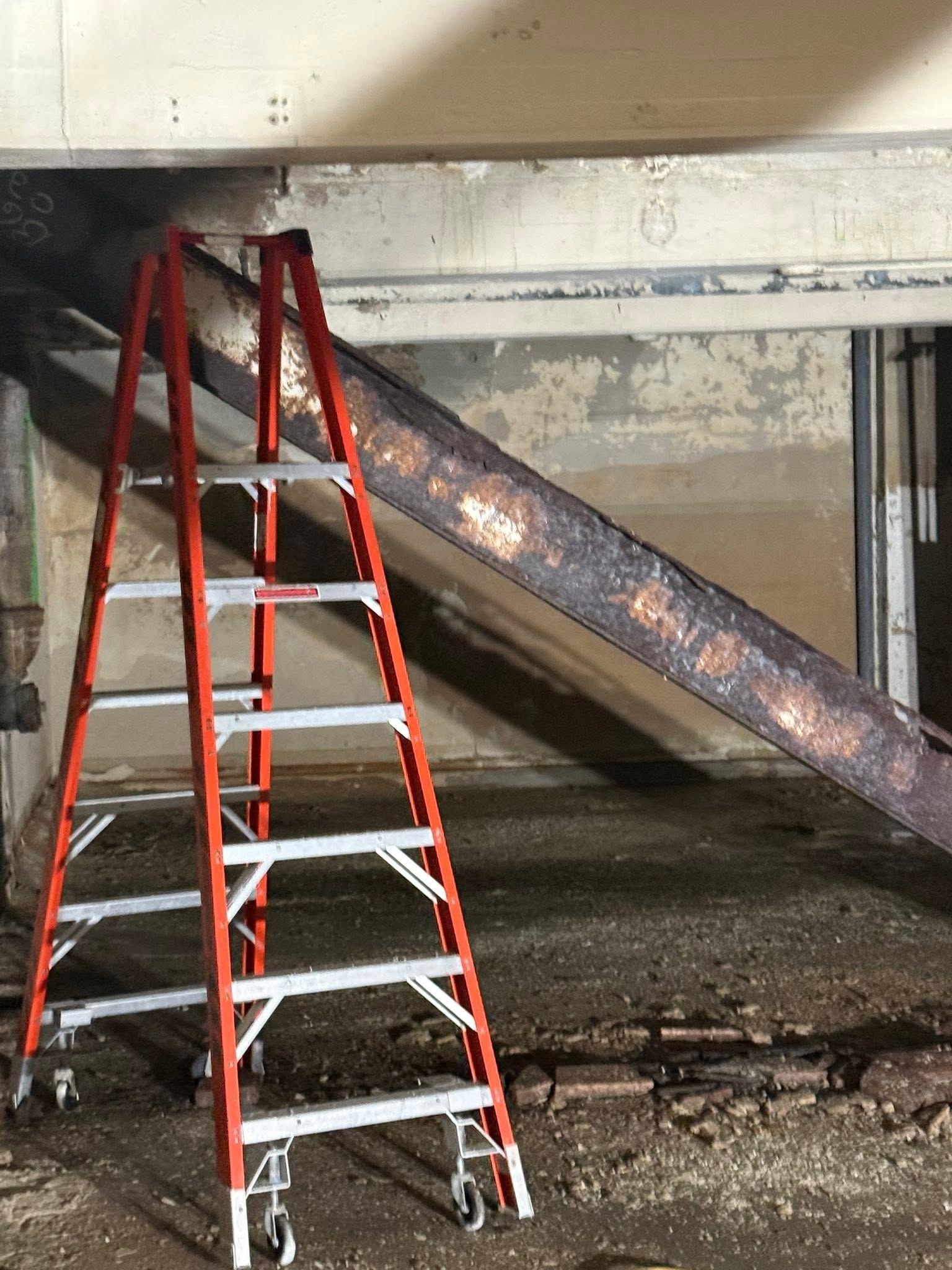 Red ladder beside a rusty diagonal steel beam in a concrete basement or crawlspace.