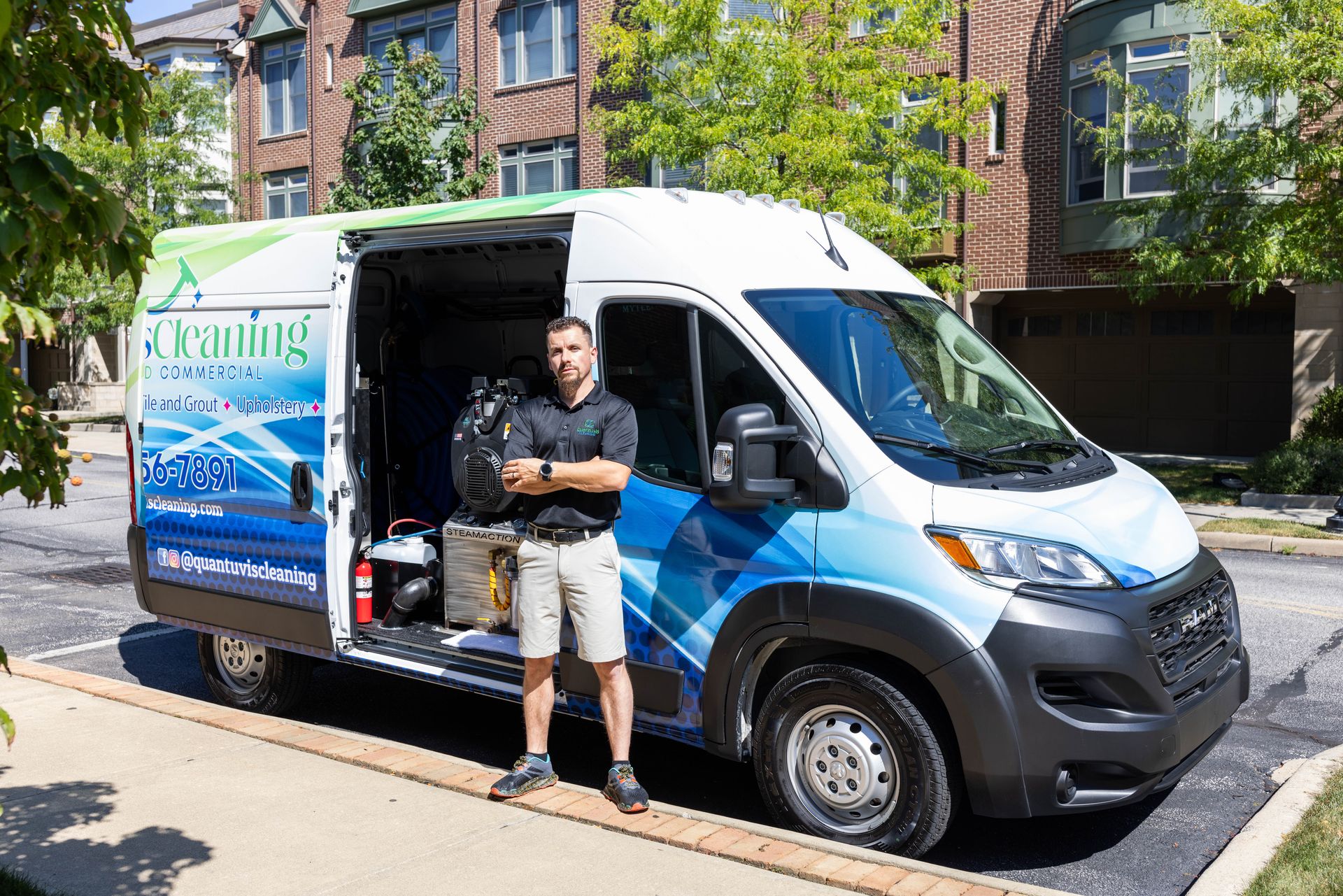 Man stands by cleaning van, arms crossed. Van is white with blue graphics. Outdoors, sunny day.