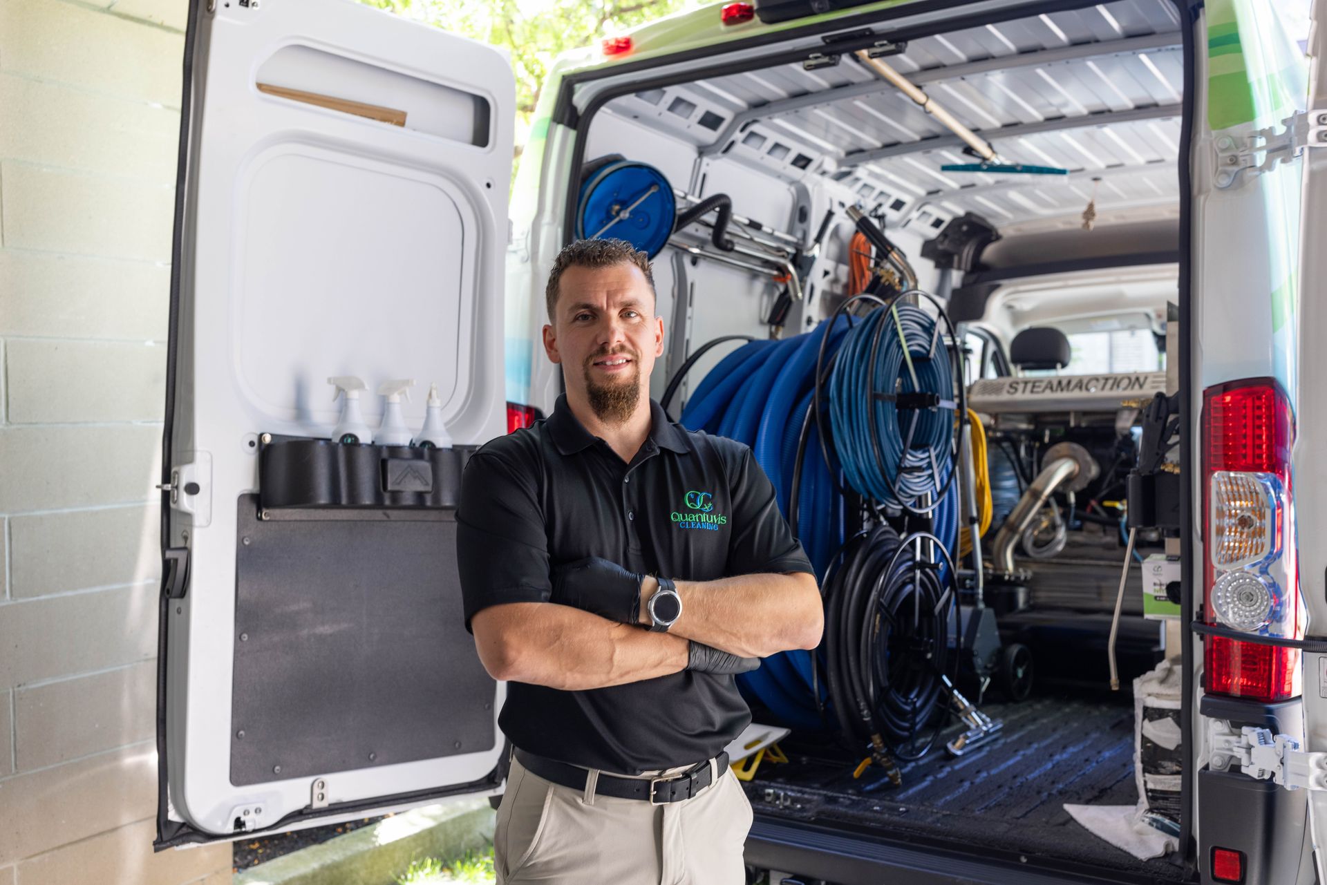 Man with arms crossed stands in open van filled with cleaning equipment.