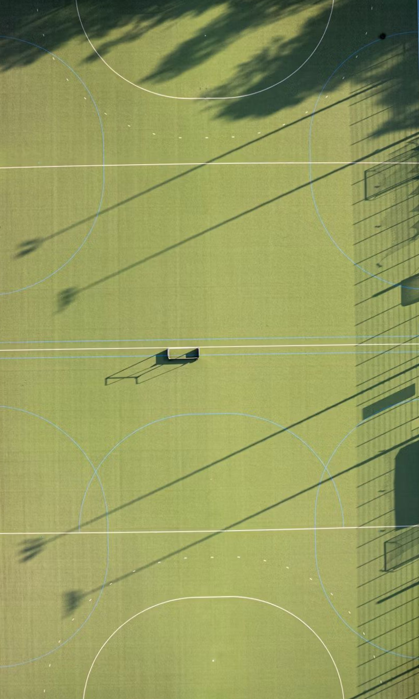 Overhead view of a green hockey field with painted white lines, shadows cast by trees, and a bench.