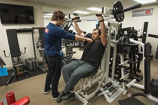 A man is sitting on a bench in a gym while a man stands behind him.