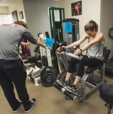 A man is standing next to a woman sitting on a machine in a gym.