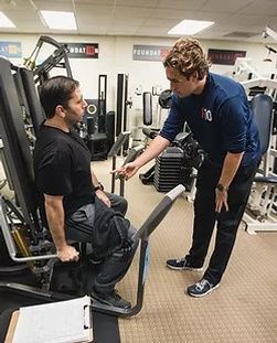 A man is sitting on a machine in a gym talking to another man.
