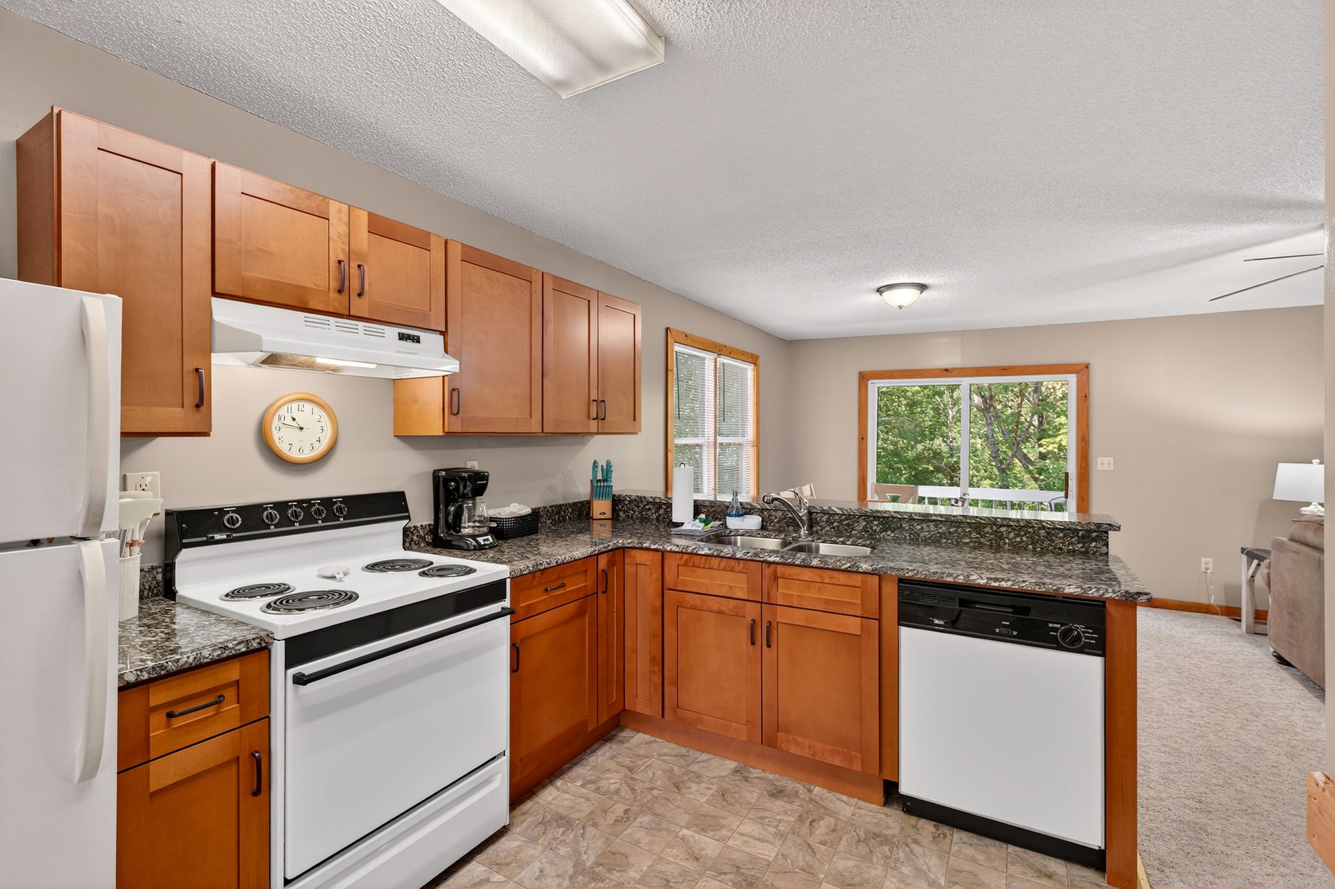 Kitchen with white appliances, wooden cabinets, granite countertops, and a window overlooking trees.