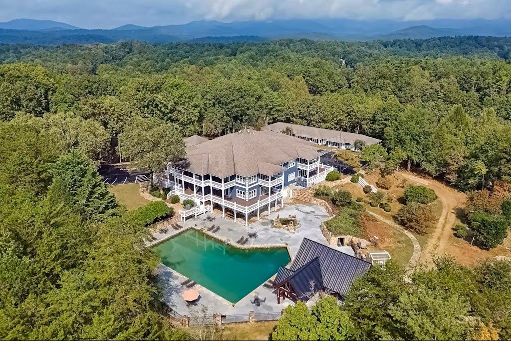 Large white mansion with pool in a lush green forest, mountains in the background.