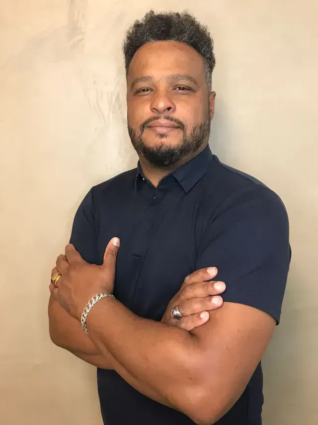 Um homem com barba está de pé com os braços cruzados.
