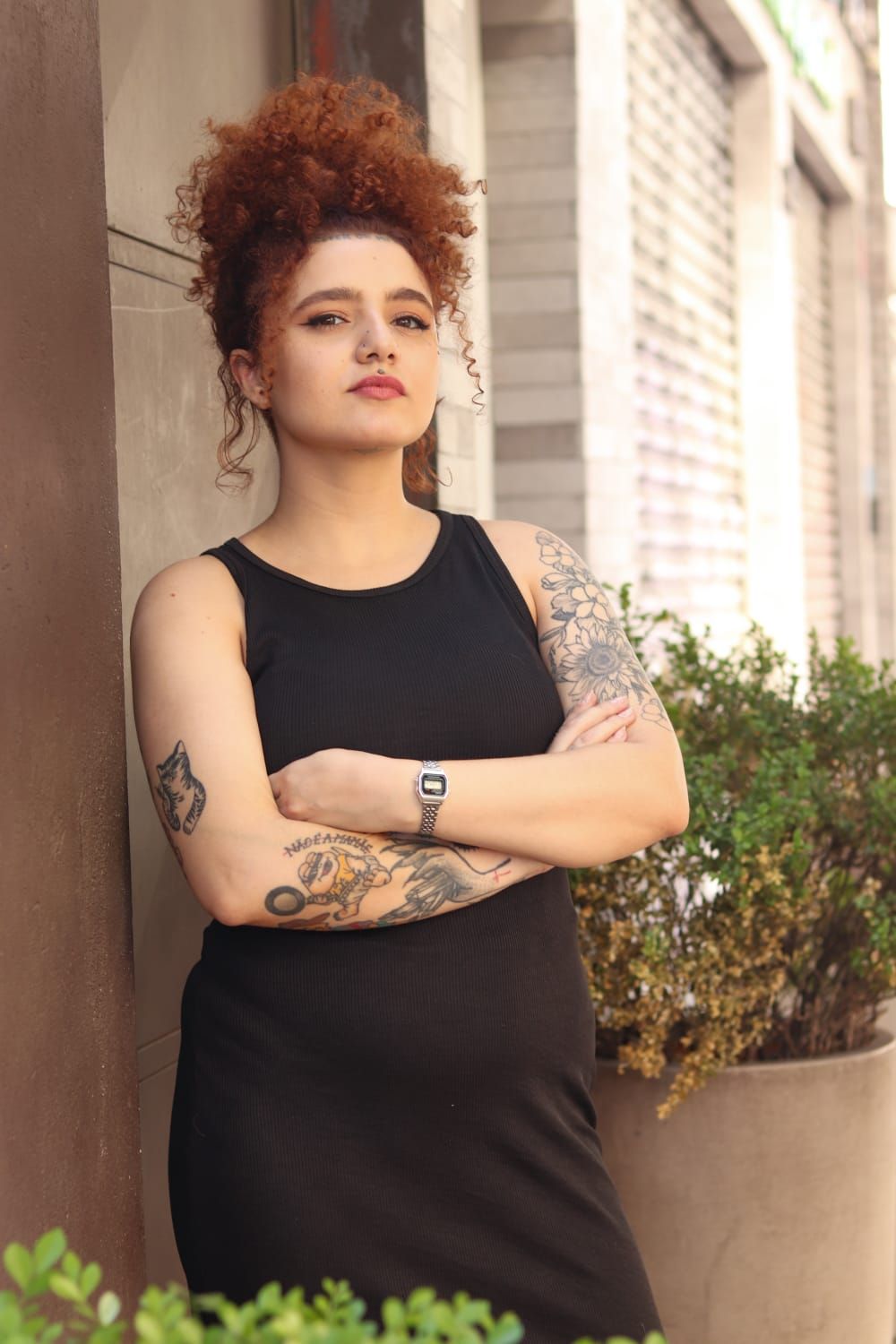 Uma mulher com tatuagens nos braços está parada em frente a um prédio com os braços cruzados.
