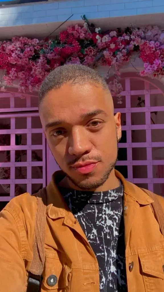 Um homem de jaqueta marrom está tirando uma selfie em frente a uma parede rosa com flores.