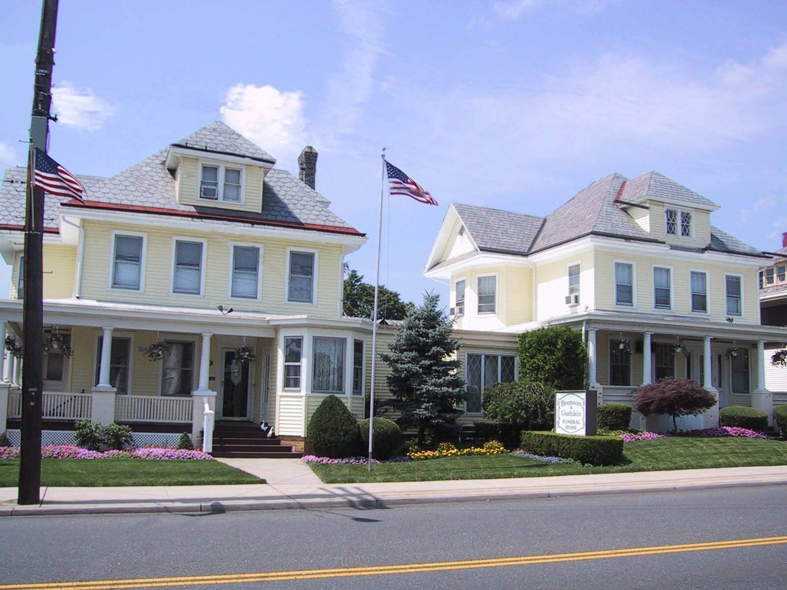 Two pale yellow houses with porches and American flags along a street under a blue sky