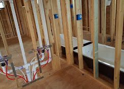Construction site showing exposed wooden wall studs, white plumbing pipes, red PEX tubing, and white bathtubs installed.