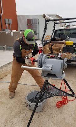 A worker in tan overalls uses a powered pipe-threading machine on a tripod stand at an outdoor job site.