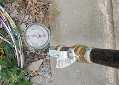 A circular pressure gauge connected to a metal pipe with a shut-off valve, situated on gravel ground next to loose cables.