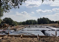 A construction site with a foundation prepared with plastic vapor barrier sheeting, wooden forms, and a tractor nearby.