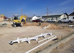 A construction site with a yellow excavator, white plumbing pipes in the foreground, and trucks parked nearby.