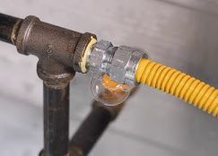 A yellow corrugated gas line fitting connected to a black iron pipe tee, with soap bubbles indicating a gas leak.