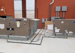 Two tan-colored York HVAC rooftop units connected by black gas piping against a red-brown building wall.