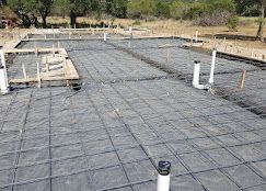Construction site showing a residential concrete slab foundation with wire mesh, vapor barrier, and upright plumbing pipes.