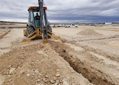 A yellow backhoe digs a long, narrow trench in a flat, dusty construction site under a cloudy sky.