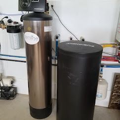A stainless steel water filtration tank and a black brine tank installed in a utility room with connected piping.