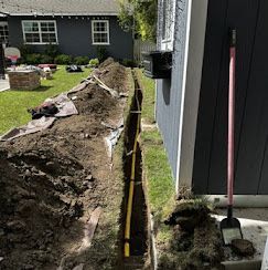 A narrow trench dug along the side of a blue house with a yellow gas line visible inside, and a shovel leaning nearby.