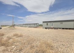 A row of long, gray modular buildings sits on a gravel lot under a bright, cloudy sky.