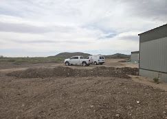 A white pickup truck and a white van parked on a dirt lot near a building under a cloudy sky.
