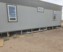 Exterior view of a light grey modular home on pillars with exposed white plumbing pipes running along the foundation.
