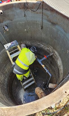 A worker in a high-visibility yellow jacket stands on a ladder inside a deep, circular concrete maintenance shaft.