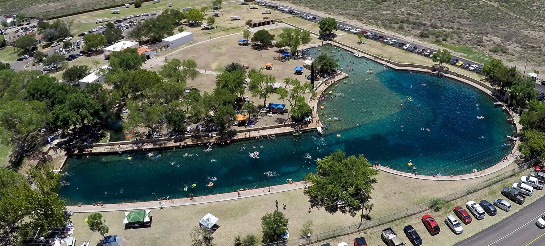Aerial view of a bright blue spring-fed pool surrounded by grassy parkland, trees, and parked cars.