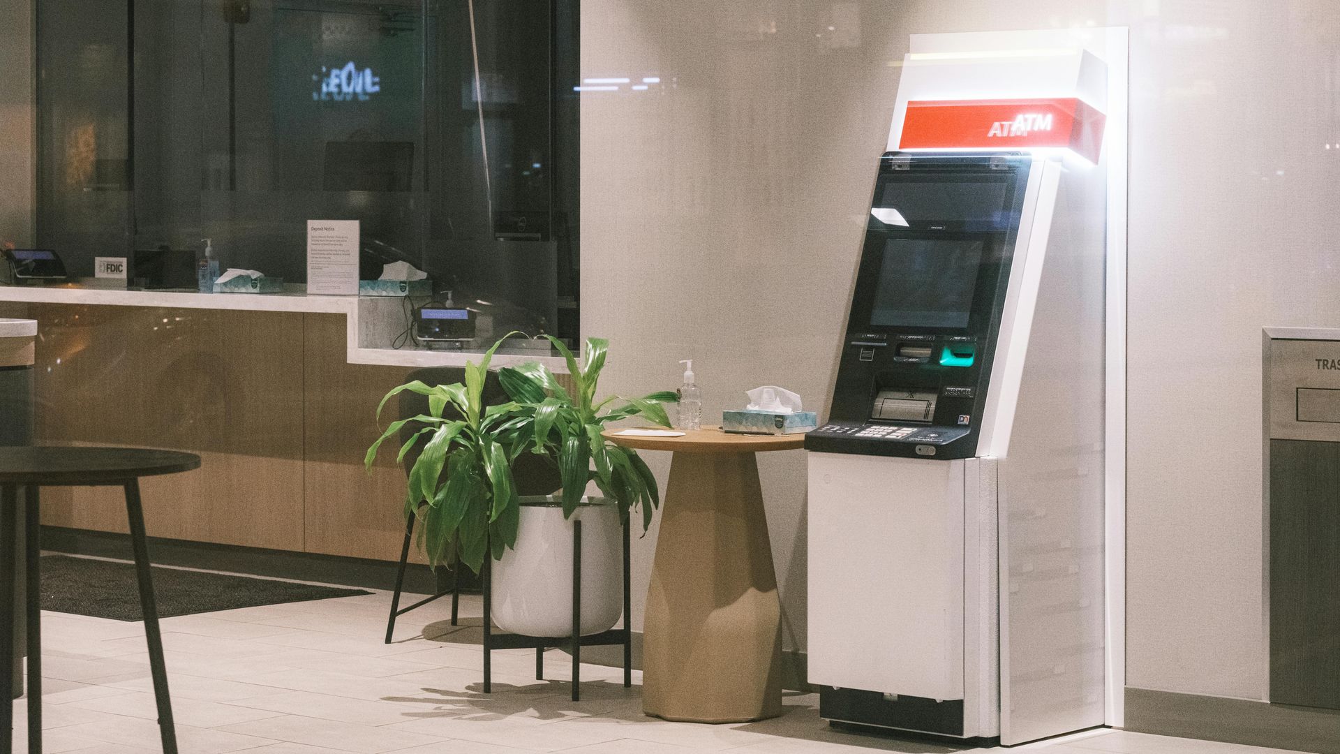 An ATM stands next to a small wooden table holding a plant and tissues in a brightly lit indoor space.