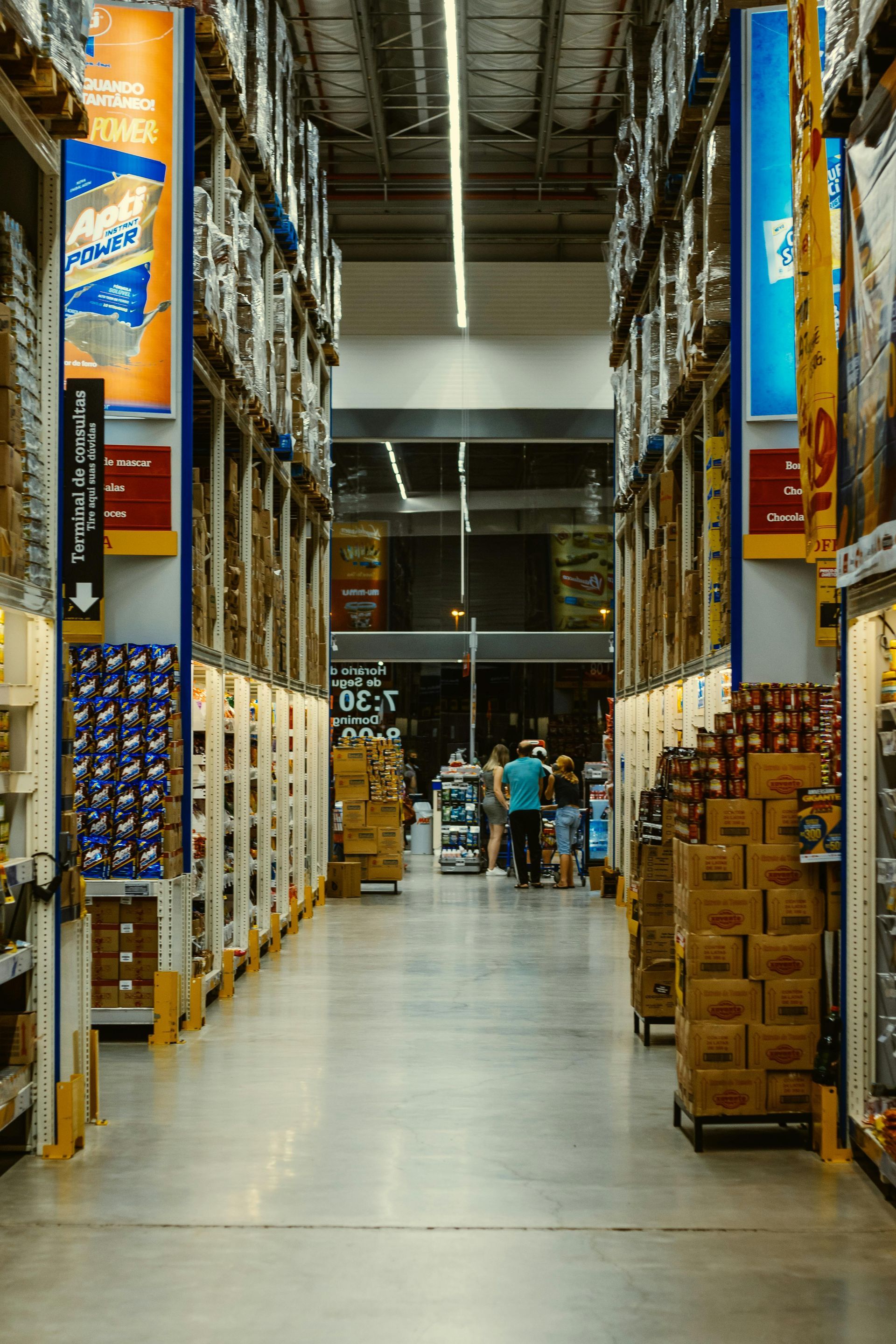 A view down a brightly lit warehouse store aisle with towering shelves of stacked merchandise and people in the distance.