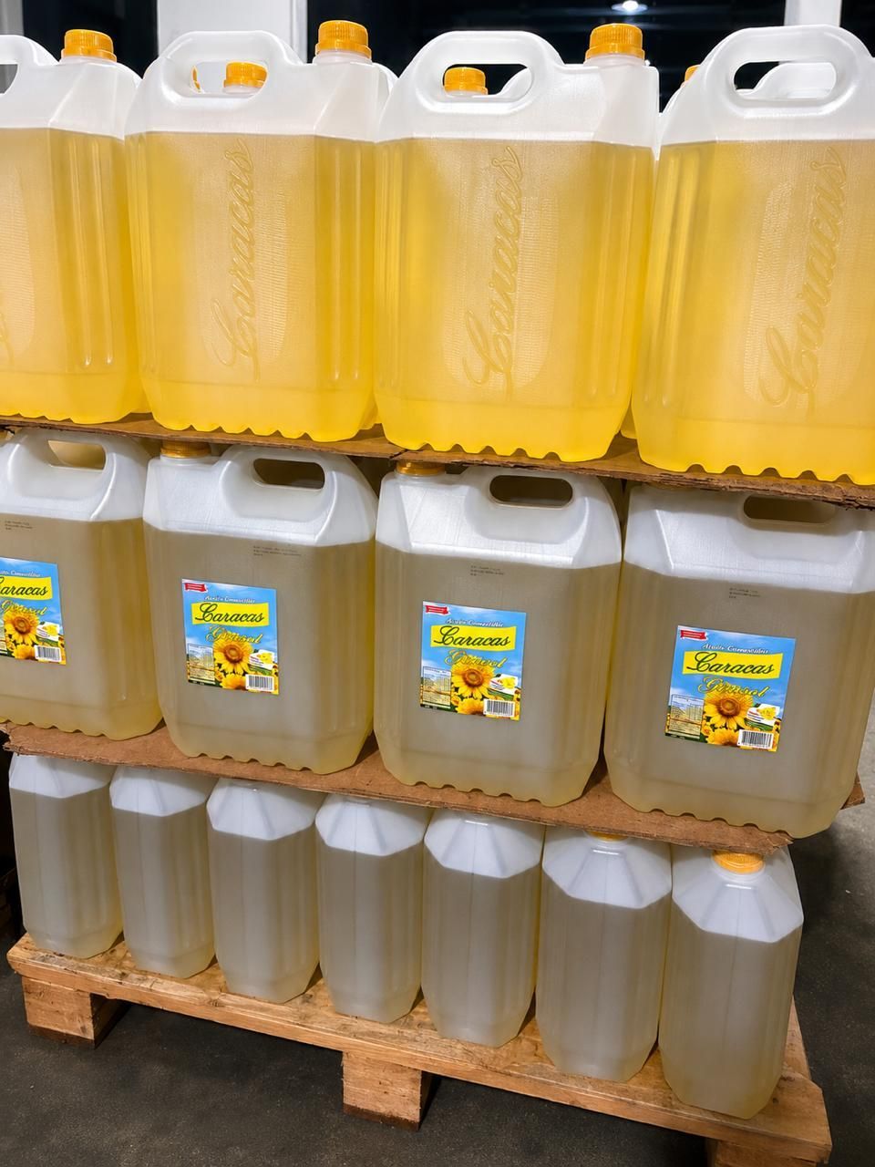 Filas de grandes garrafas de plástico llenas de líquido amarillo y marrón sobre palés en un pasillo de un almacén.