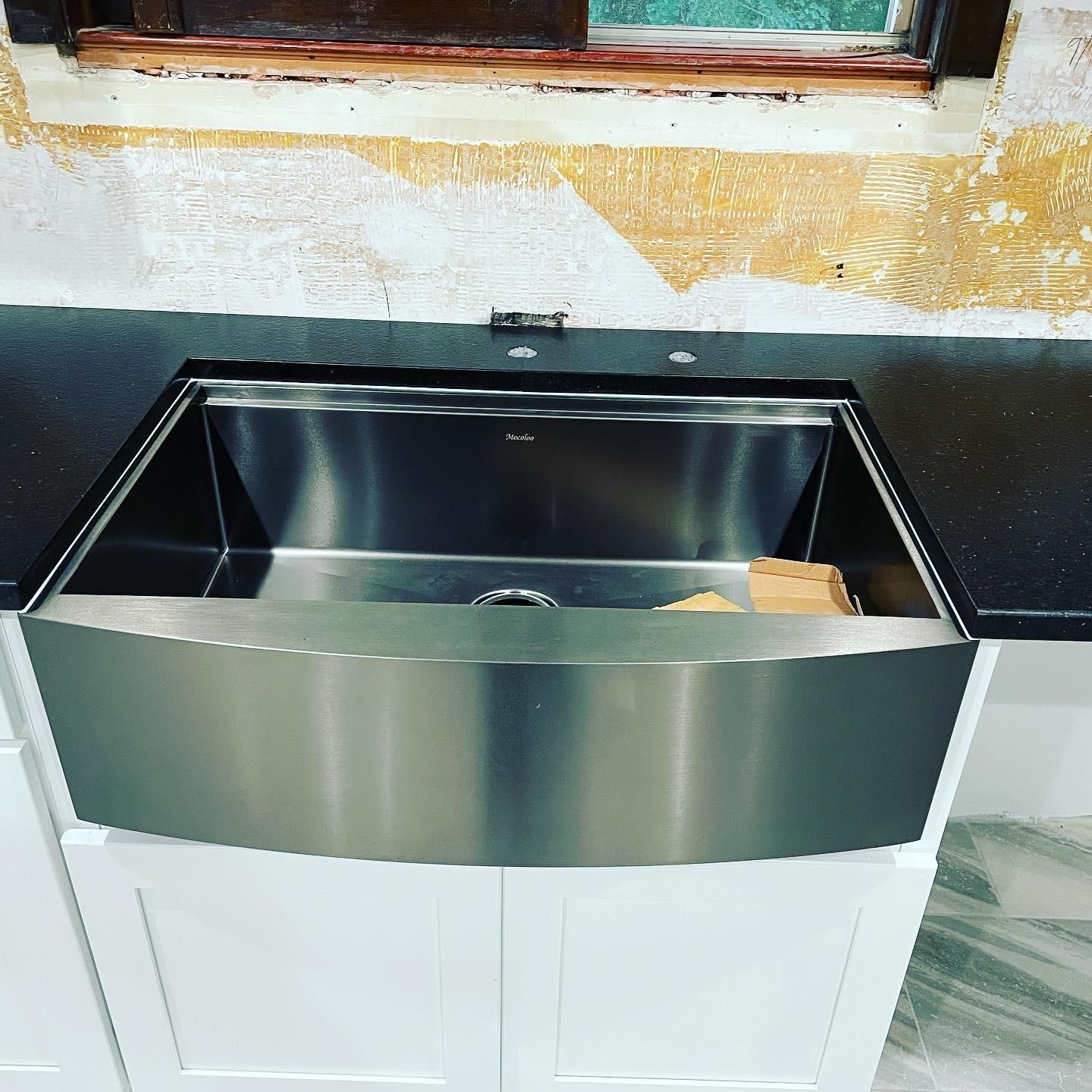 Stainless steel farmhouse sink installed in a black countertop, above white cabinets.